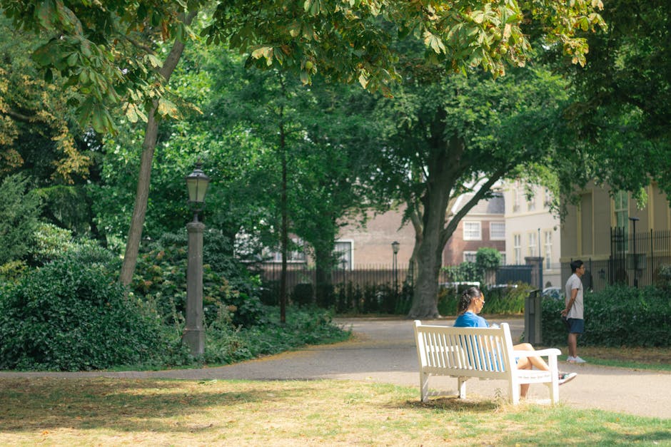 A woman sitting on a white park bench, wearing a blue shirt and beige shorts, is visible in a green outdoor area with tall trees, shrubs, and a paved pathway. Nearby, a young girl with a ponytail, dressed in a blue jacket and white skirt, is seated with her legs crossed on the bench, looking towards the right. An older boy, wearing a white T-shirt and dark shorts, stands on the pathway facing away from the camera. To the left of the scene, there is a black lamp post among the foliage, and the background features residential buildings with white facades and black fences. The scene is well-lit with natural sunlight, highlighting the lush greenery and the various objects involved in a typical house removal or home relocation process. Although the image primarily depicts a peaceful outdoor setting, it subtly reflects the process of packing and moving, with the outdoor environment serving as a representation of a home being prepared for transport and relocation, similar to services offered by Holland Park Man and Van.