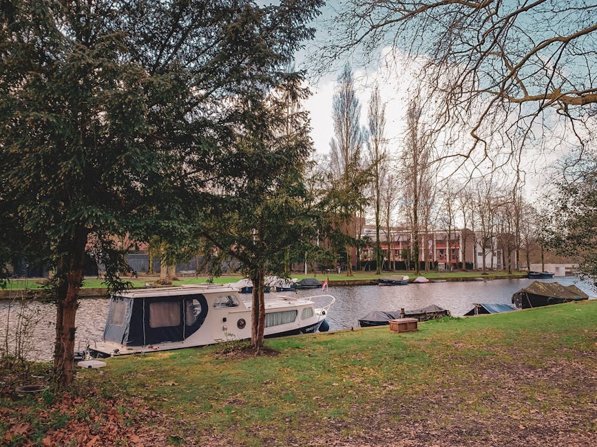 A houseboat moored along the edge of a narrow river in a residential area with modern multi-storey buildings visible in the background. The houseboat has a white exterior with a dark blue canvas canopy covering the entrance, and it is positioned close to the grassy bank, partly hidden behind trees with bare branches and dense foliage. Several other boats, some covered with tarps, are also tied along the riverbank. The scene appears to be during late autumn or early winter, with leafless trees and subdued lighting. In the foreground, the grassy area includes a small wooden crate, and the overall environment suggests a peaceful waterside location suitable for a home relocation or furniture transport project, potentially managed by Holland Park Man and Van as part of their removals services.