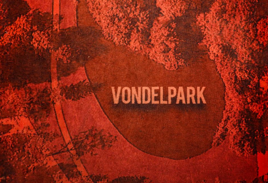 Aerial view of Vondelpark in Amsterdam, featuring a large oval-shaped lawn area surrounded by dense trees with reddish-orange foliage, possibly during autumn. A paved pathway runs alongside the park, with a few small pedestrians visible on the walkway. The park's name, 'VONDEL PARK,' is prominently displayed in large white capital letters within the grassy area. The scene is captured in warm, natural daylight, highlighting the contrast between the lush greenery and the vibrant tree colors. This image is relevant to house removals, as it depicts urban green spaces often encountered during residential relocations in the area, aligning with the context of furniture transport and home relocation logistics with [COMPANY_NAME] at ademandes.biz, who handle removals within this environment.