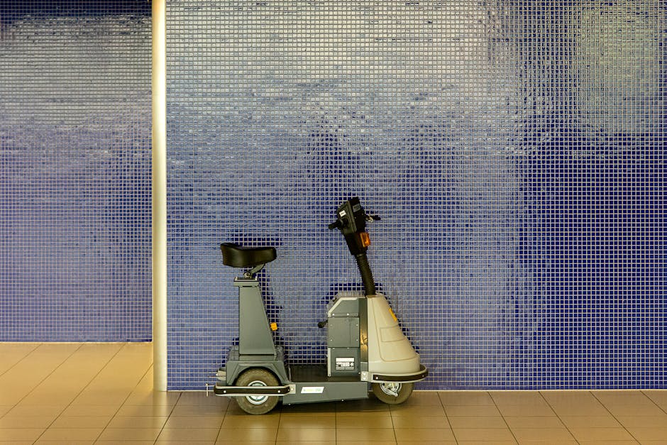 A compact indoor moving trolley with a black padded seat, a control handle, and a silver base is positioned on a beige tiled floor against a blue mosaic-tiled wall. The trolley is designed for facilitating the transport of small furniture or boxes during a home relocation. To the left of the trolley, a white vertical pole is attached to the wall, possibly part of building infrastructure or safety equipment. The scene is well-lit, highlighting the details of the trolley and the textured tiles. The setting appears to be a hallway or corridor within a residential or commercial property, prepared for loading or unloading as part of a house removal service. This image exemplifies the type of equipment used by Holland Park Man and Van for efficient furniture transport and packing during the moving process, emphasizing attention to within-property access considerations for effective removals in the Holland Park area.